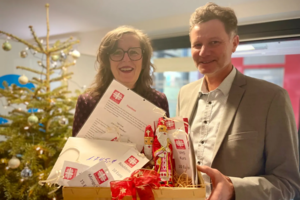 Zwei Personen halten einen Geschenkkorb mit verpackten Lebensmitteln und einem Brief, im Hintergrund ein geschmückter Weihnachtsbaum