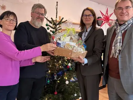 Vier Personen übergeben vor einem geschmückten Weihnachtsbaum einen Geschenkkorb