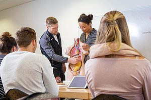Ein Dozent zeigt auf ein anatomisches Modell des menschlichen Körpers. Im Vordergrund sind mehrere Studierende zu sehen, die auf den Vortrag achten.