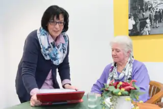Eine jüngere Frau zeigt einer älteren Frau an einem Tisch etwas auf einem roten Tablet, daneben steht eine Vase mit Blumen.
