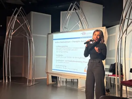 Frau steht mit Mikrofon vor einer Leinwand mit Präsentationsfolie in einem Raum mit architektonischen Bögen