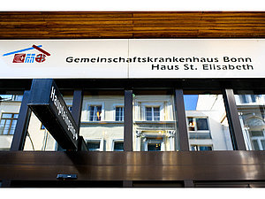 Eingangsbereich mit Schild 'Gemeinschaftskrankenhaus Bonn Haus St. Elisabeth' und schwarzem Wegweiser 'Haupteingang' vor Glasfassade eines Gebäudes