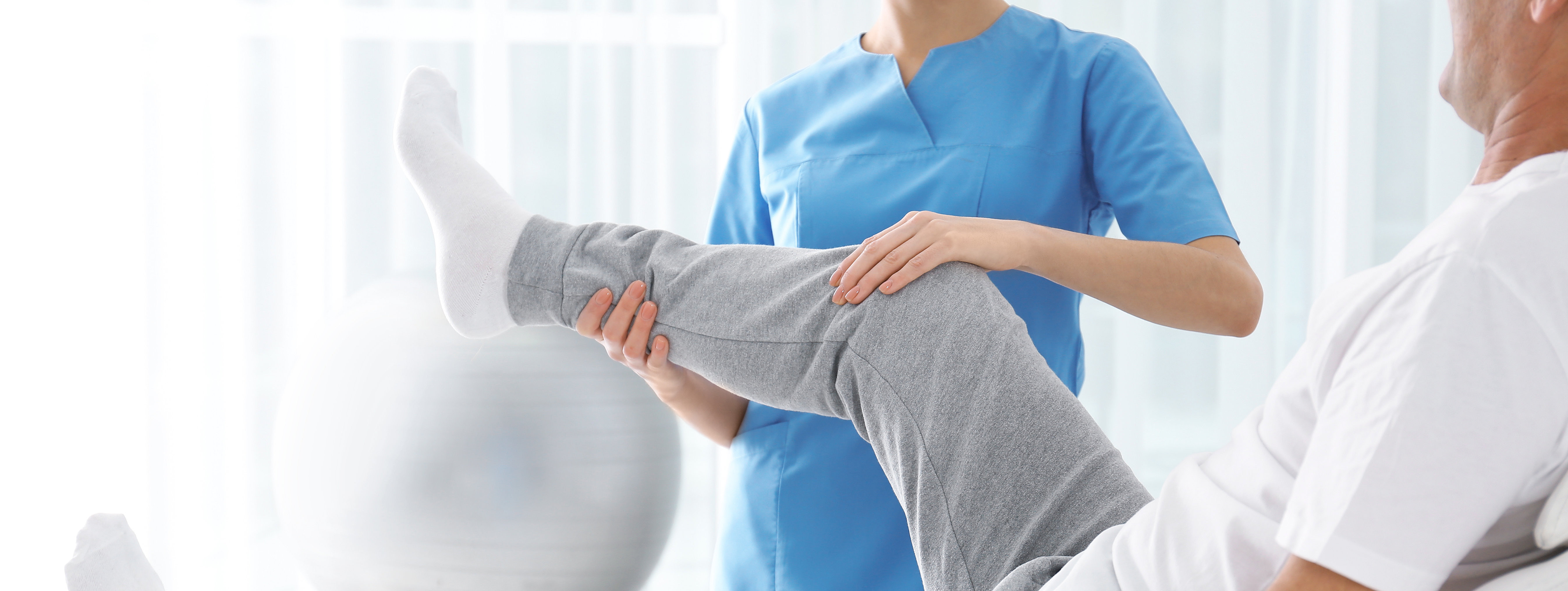 Physiotherapeutin in blauer Kleidung hebt das angewinkelte Bein eines älteren Mannes auf einer Liege.