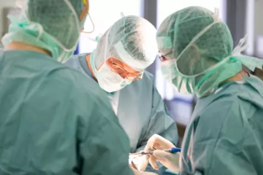 Drei Chirurgen in grüner OP-Kleidung und Schutzmasken bei einer Operation im OP-Saal.