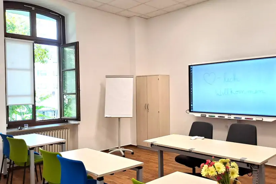 Klassenzimmer mit mehreren Tischen und bunten Stühlen, Flipchart, Schrank und großem Bildschirm mit handgeschriebener Willkommensnachricht