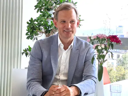 Mann in hellblauem Sakko und weißem Hemd sitzt an einem Tisch mit Blumen und Pflanzen im Hintergrund