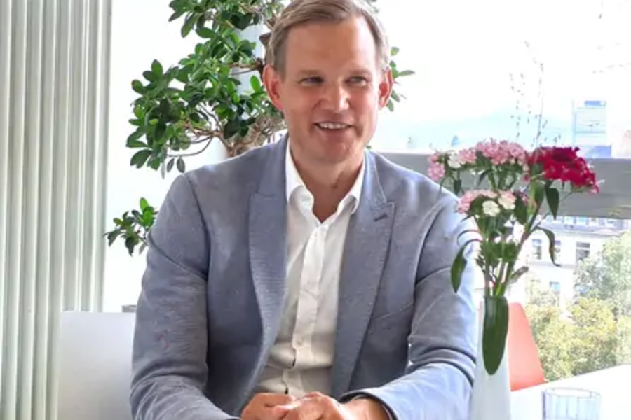 Mann in hellblauem Sakko und weißem Hemd sitzt an einem Tisch mit Blumen und Pflanzen im Hintergrund
