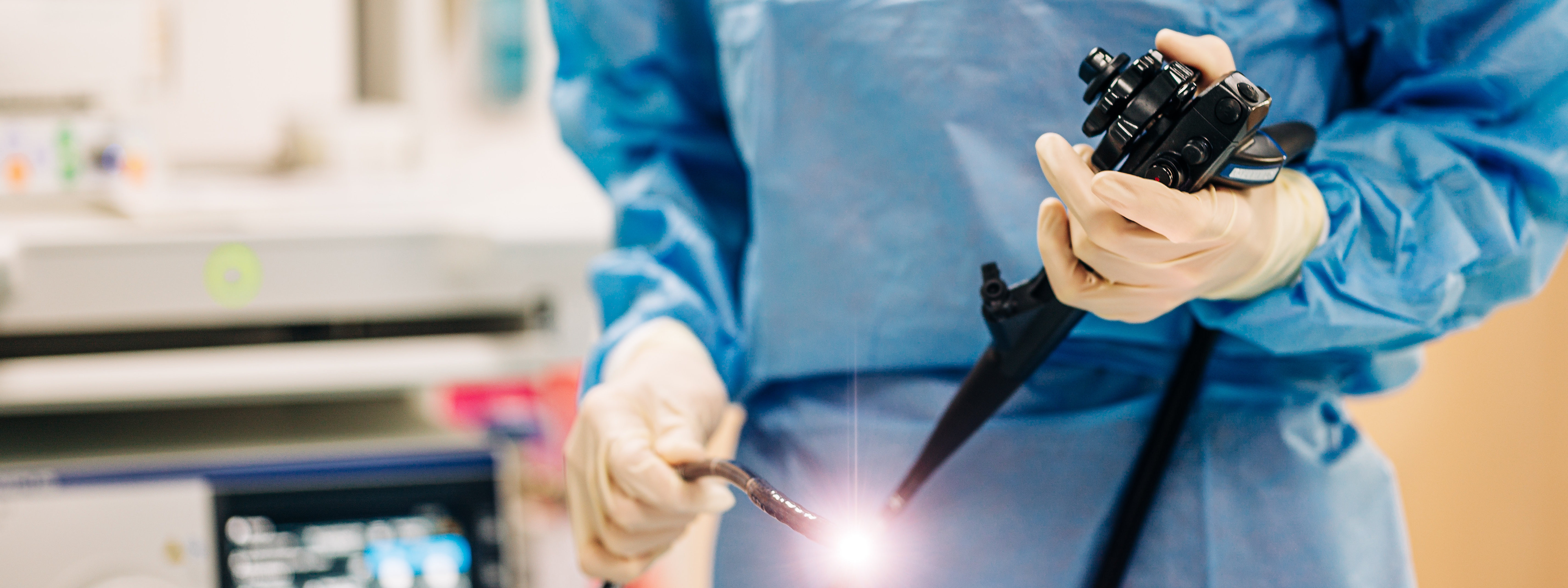 Eine Person in blauer Schutzkleidung hält ein medizinisches Endoskop und eine Lichtquelle in der Hand. Im Hintergrund ist ein medizinisches Gerät sichtbar.