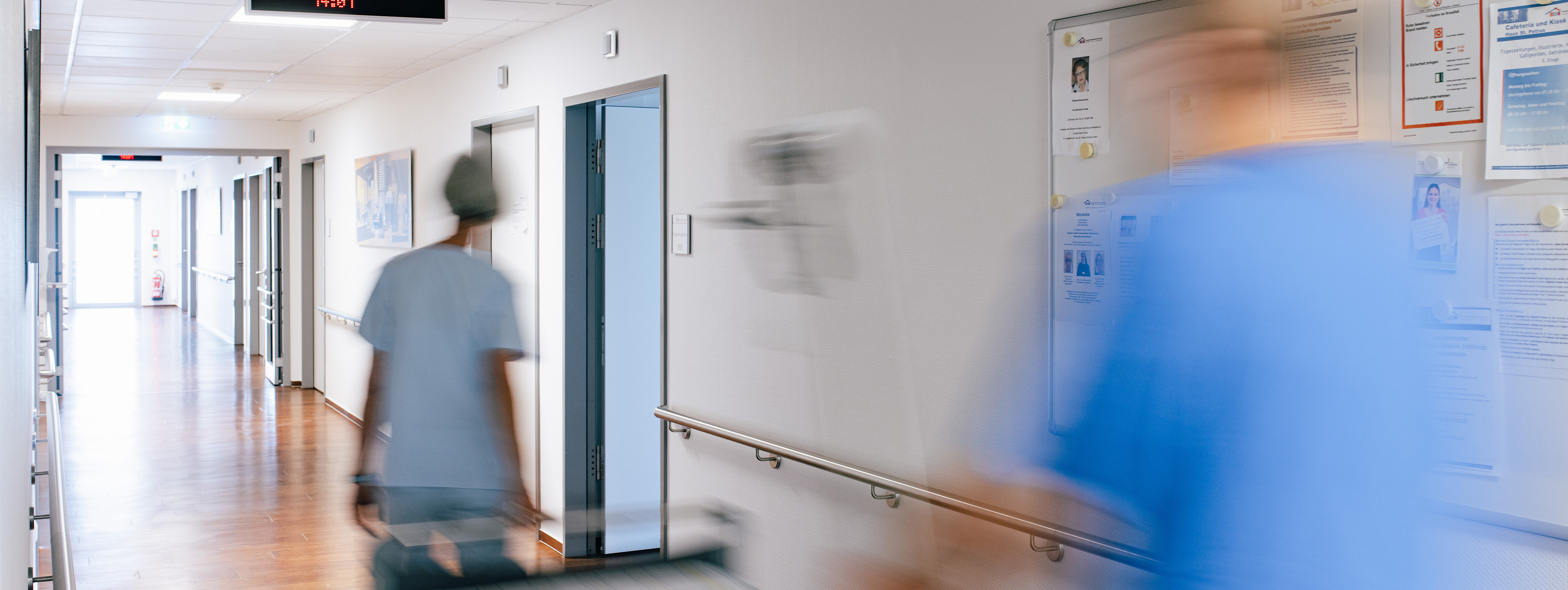 Unscharfe Aufnahme eines Krankenhausflurs mit zwei Personen in medizinischer Kleidung, eine schiebt ein Bett.