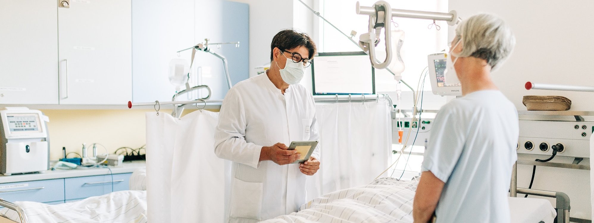 Krankenhauszimmer mit einem Arzt in weißem Kittel, der ein Tablet hält, und einer Patientin im hellen Nachthemd am Bett