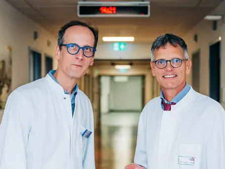 Zwei Männer in weißen Kitteln stehen in einem Krankenhausflur mit beleuchtetem Notausgangsschild im Hintergrund