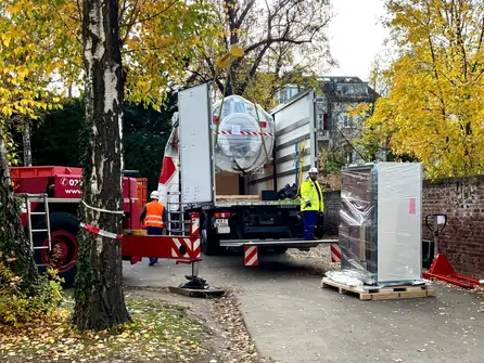 LKW mit geöffnetem Heck und Hebebühne lädt verpackte Geräte auf einem Gehweg, umgeben von Bäumen mit herbstlichem Laub und zwei Arbeitern in Warnkleidung