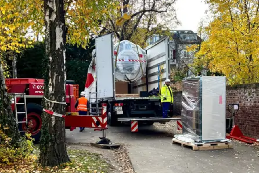 LKW mit geöffnetem Heck und Hebebühne lädt verpackte Geräte auf einem Gehweg, umgeben von Bäumen mit herbstlichem Laub und zwei Arbeitern in Warnkleidung