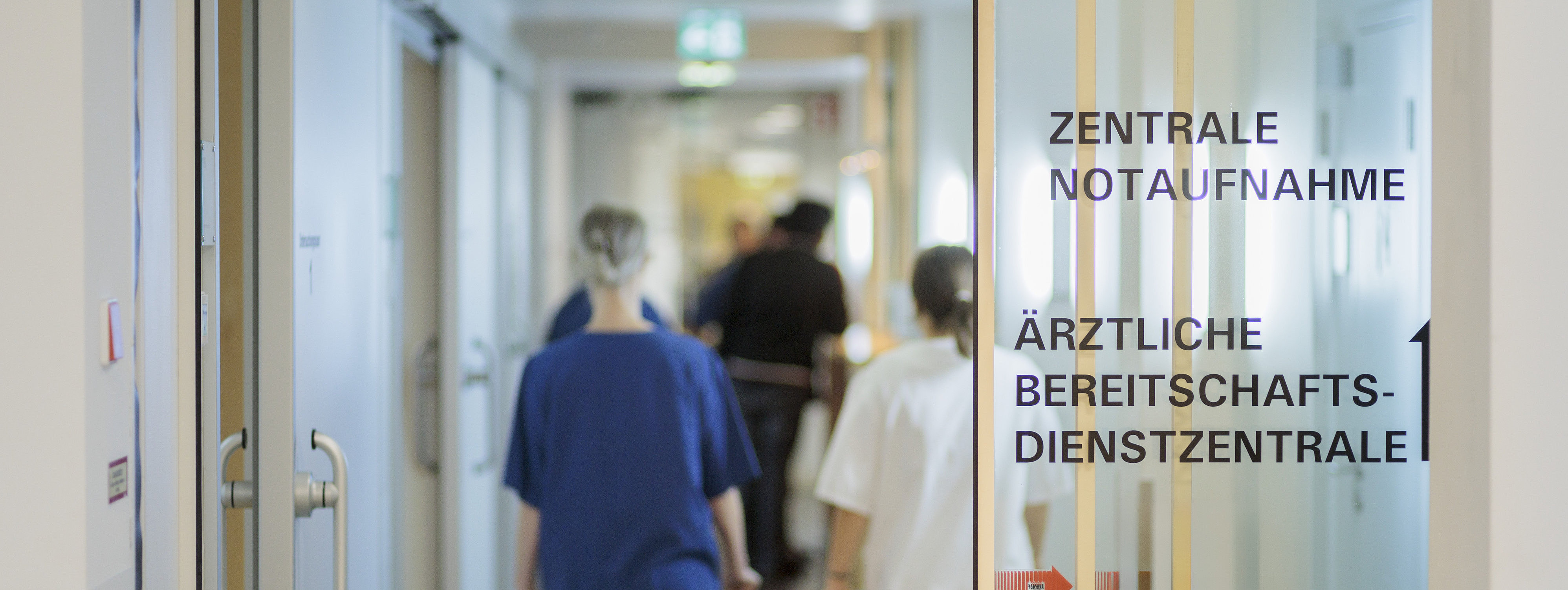 Krankenhausflur mit mehreren Personen in medizinischer Kleidung, rechts an der Wand steht 'ZENTRALE NOTAUFNAHME' und 'ÄRZTLICHE BEREITSCHAFTSDIENSTZENTRALE'.