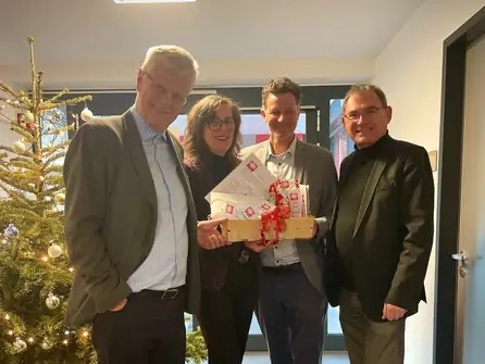 Vier Personen stehen in einem Raum mit Weihnachtsbaum, eine Person hält ein Geschenkpaket mit roten Schleifen und Briefen