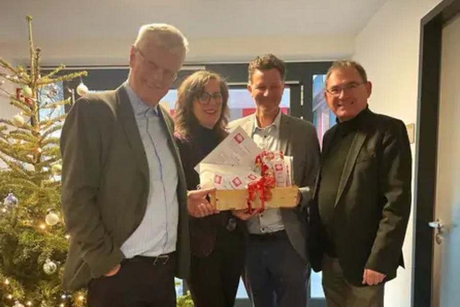 Vier Personen stehen in einem Raum mit Weihnachtsbaum, eine Person hält ein Geschenkpaket mit roten Schleifen und Briefen