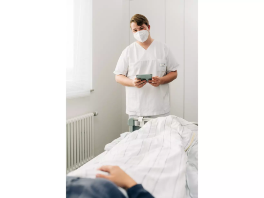 Krankenpfleger in weißer Arbeitskleidung steht an einem Bett und hält ein Tablet