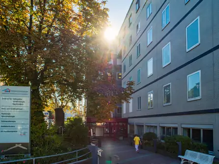 Eingang eines mehrstöckigen Gebäudes mit Person in gelber Kleidung, Bänken und Bäumen bei Sonnenlicht