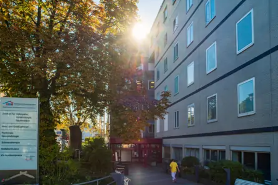 Eingang eines mehrstöckigen Gebäudes mit Person in gelber Kleidung, Bänken und Bäumen bei Sonnenlicht