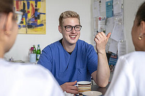 Ein Mann in blauer Arbeitskleidung sitzt an einem Tisch und gestikuliert mit der rechten Hand. Zwei Personen in weißen T-Shirts sitzen ihm gegenüber.