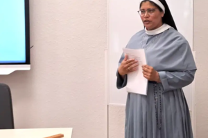 Nonne in grauem Habit hält Papiere und steht in einem Raum mit weißer Wand und einem Holzkreuz auf einem Tisch