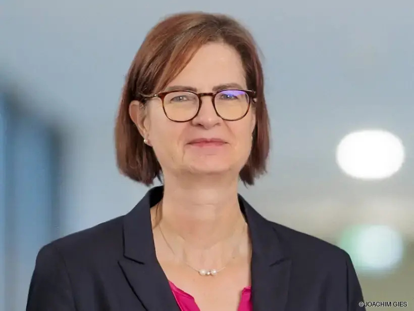 Frau mit schulterlangen braunen Haaren trägt schwarzen Blazer und pinkes Oberteil, steht vor unscharfem Hintergrund