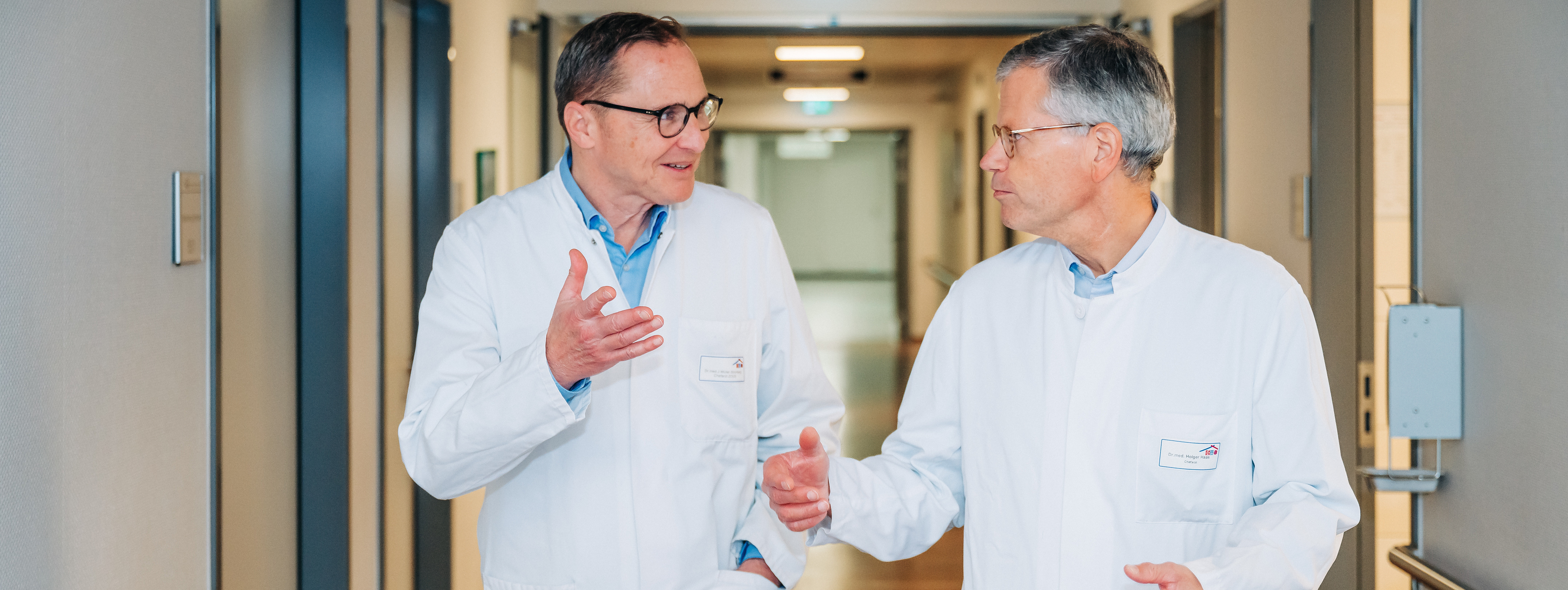Zwei Männer in weißen Arztkitteln gehen und unterhalten sich in einem Krankenhausflur.