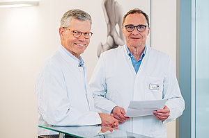 Zwei Männer in weißen Kitteln stehen nebeneinander, einer hält ein Blatt Papier
