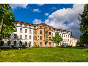 Großes historisches Gebäude mit mehreren Fenstern und einem verzierten Mittelteil vor einer Wiese mit Bäumen und blauem Himmel mit Wolken