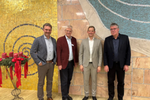 Vier Männer in Anzügen stehen vor einer Wand mit gelben und blauen Mosaiken, links ein Ständer mit roten Kerzen und Bändern