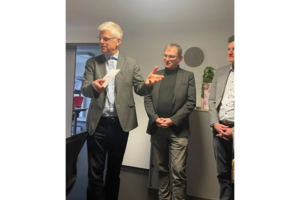 Drei Männer in Geschäftskleidung stehen in einem Büro, einer spricht und gestikuliert, die anderen hören zu