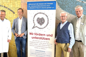 Vier Männer stehen neben einem Banner der Gesellschaft der Freunde und Förderer des Gemeinschaftskrankenhauses Bonn mit dem Text 'Wir fördern und unterstützen'.