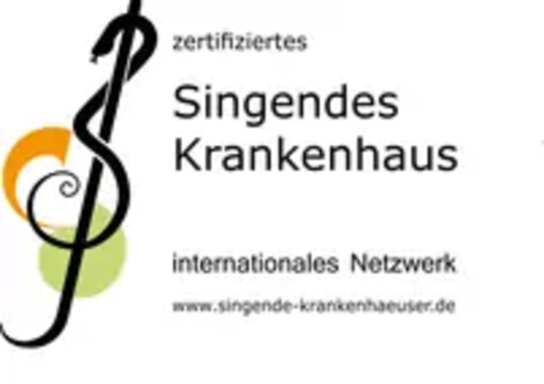 Logo mit einem stilisierten Notenschlüssel, der einen Schlangenstab darstellt, begleitet von den Worten 'zertifiziertes Singendes Krankenhaus internationales Netzwerk' und der Webadresse 'www.singende-krankenhaeuser.de'