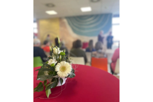 Blumenarrangement mit weißen Blüten auf rotem Tisch, unscharfer Hintergrund mit Menschen in einem Raum