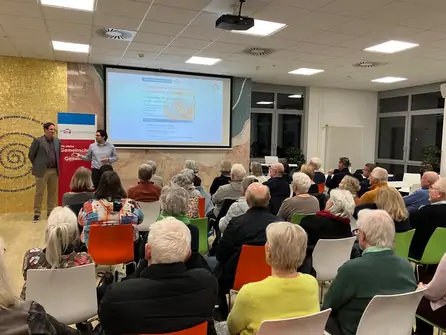 Zwei Vortragende präsentieren vor einer Gruppe älterer Menschen in einem Raum mit Projektor und Leinwand