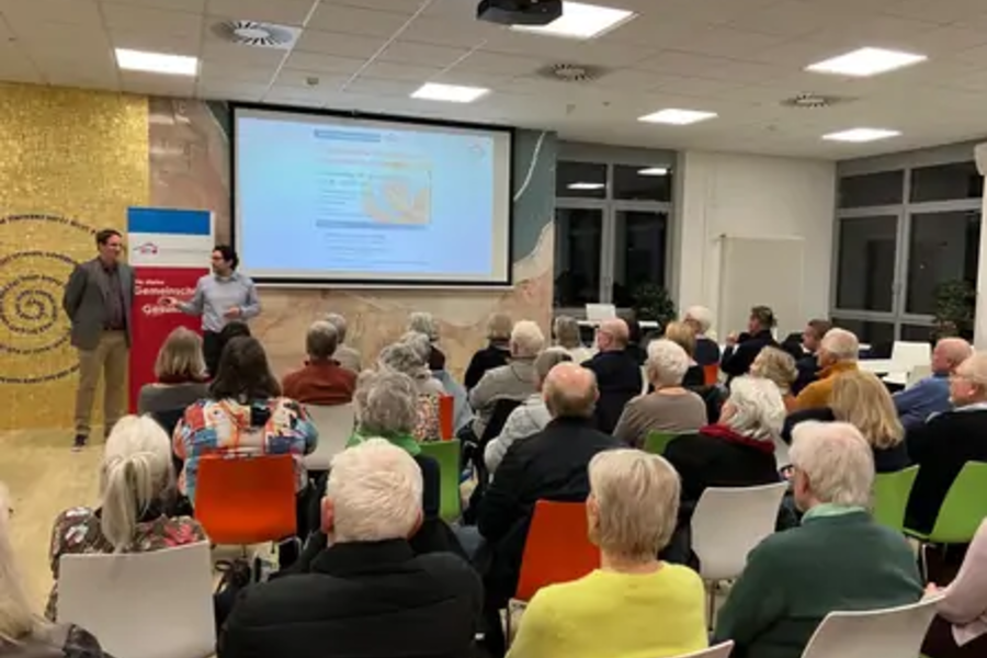 Zwei Vortragende präsentieren vor einer Gruppe älterer Menschen in einem Raum mit Projektor und Leinwand