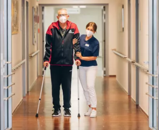 Pflegekraft stützt gehenden Mann mit Krücken im Krankenhausflur