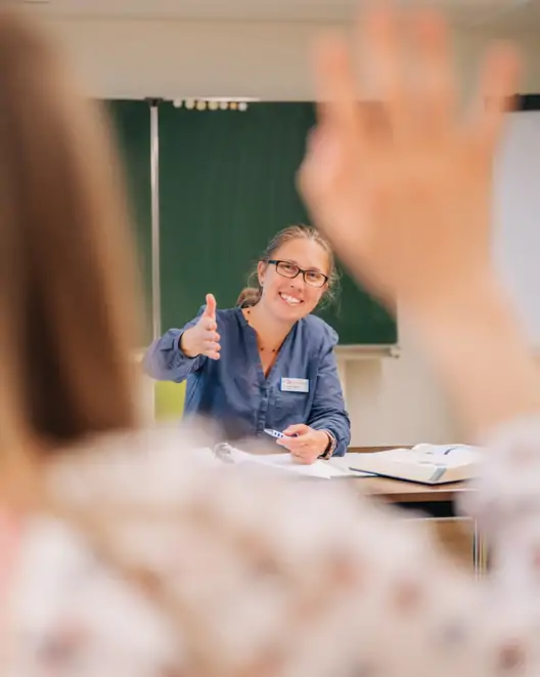 Lehrerin im Klassenzimmer zeigt Daumen hoch und blickt auf einen Schüler mit erhobener Hand