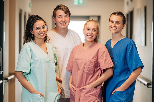 Vier Personen in Krankenhauskleidung stehen in einem Flur und blicken in die Kamera