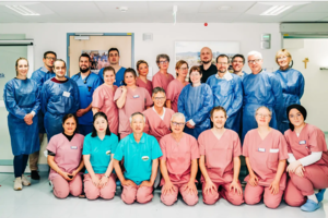Gruppenfoto von 22 medizinischen Fachkräften in Krankenhauskleidung, einige in blauen Schutzkitteln, andere in rosa oder türkisfarbener Arbeitskleidung, in einem hellen Raum