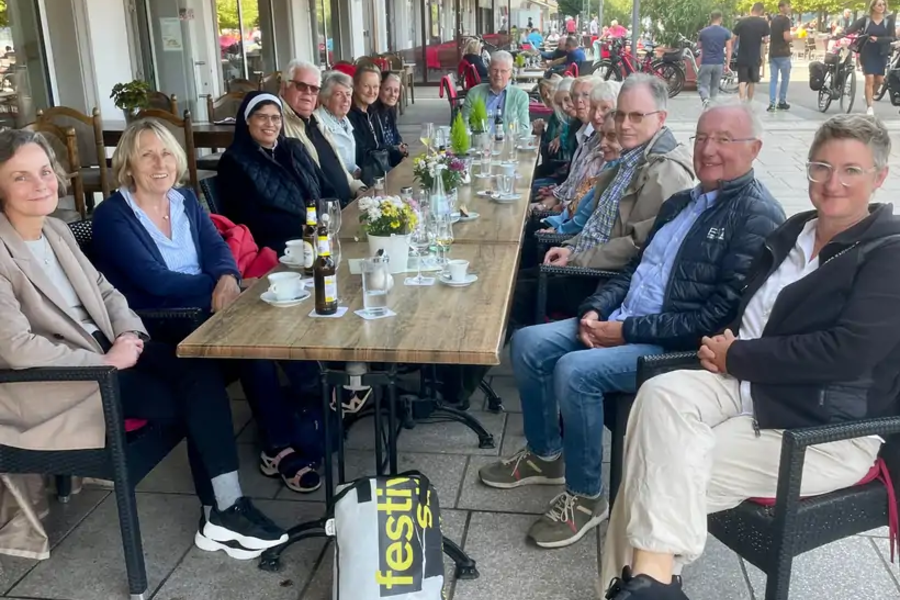 Menschen sitzen an einem langen Holztisch im Freien vor einem Gebäude mit Markise, trinken und unterhalten sich