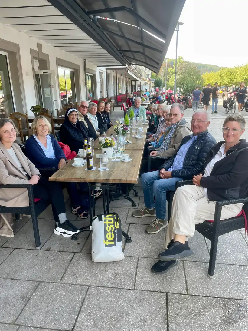Menschen sitzen an einem langen Holztisch im Freien vor einem Gebäude mit Markise, trinken und unterhalten sich