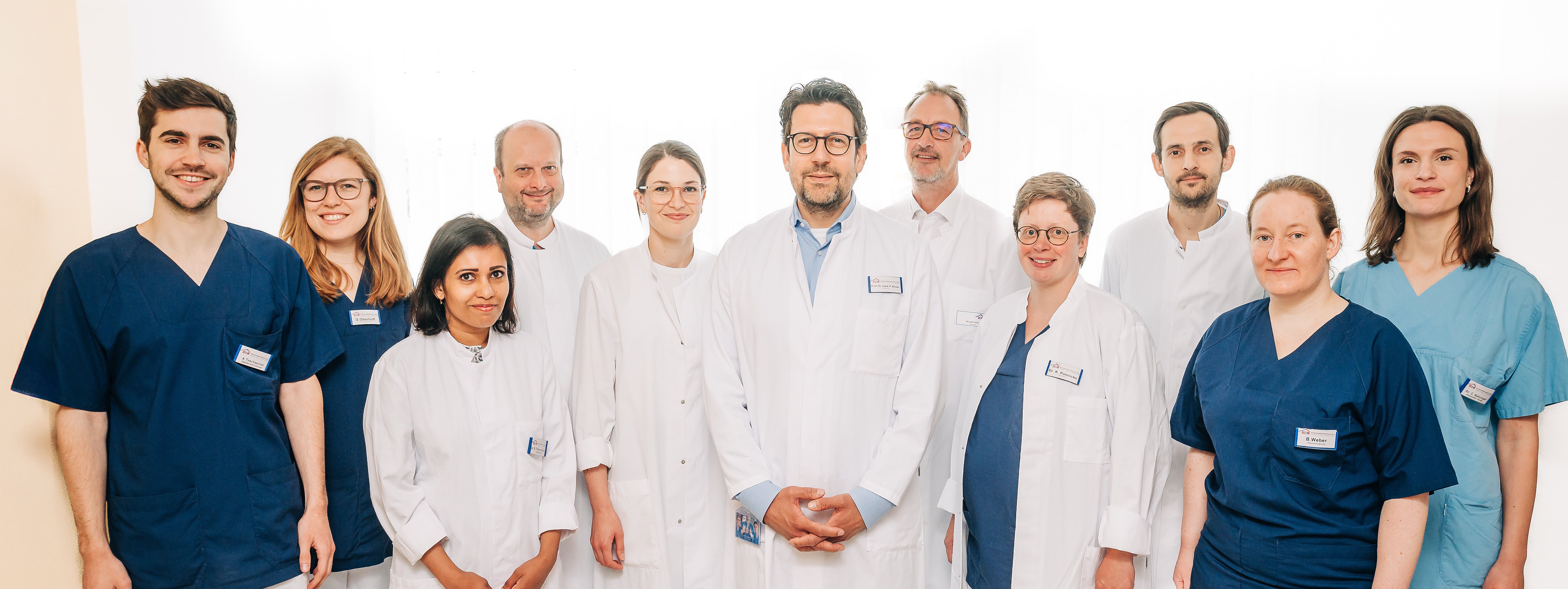 Gruppe von elf medizinischen Fachkräften in weißen Kitteln und blauen oder türkisfarbenen Kasacks, stehend in einem Flur