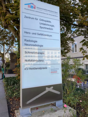 Freistehendes Informationsschild mit mehreren medizinischen Fachbereichen vor einem Gebäude und Bäumen im Hintergrund