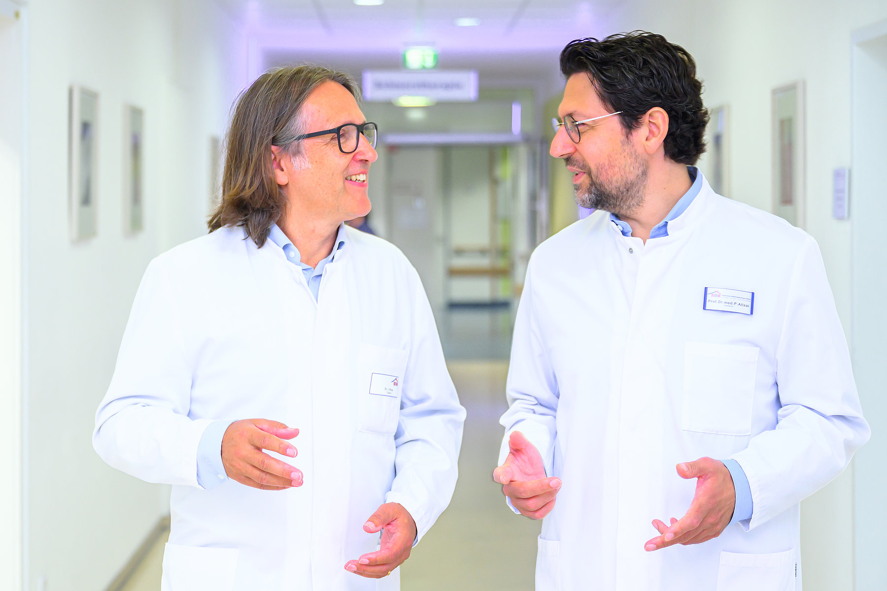 Zwei Personen in weißen Kitteln unterhalten sich in einem hellen Krankenhausflur.