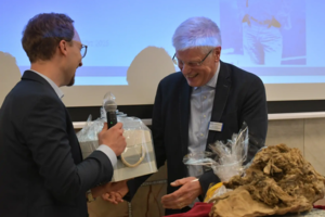 Zwei Männer in Anzügen übergeben ein verpacktes Geschenk während einer Veranstaltung