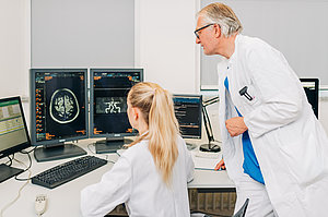 Zwei Ärzte in weißen Kitteln betrachten Gehirnscans auf mehreren Monitoren in einem medizinischen Untersuchungsraum