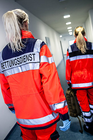 Zwei Rettungssanitäterinnen in roten Uniformen gehen einen Flur entlang, eine trägt Handschuhe und die andere zieht einen Rollkoffer