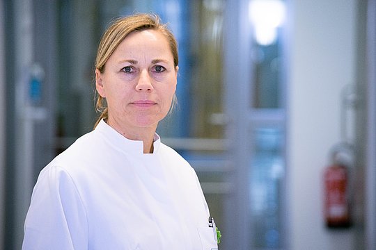 Person in weißem Arztkittel in einem hellen Flur mit Glaswänden und rotem Feuerlöscher im Hintergrund