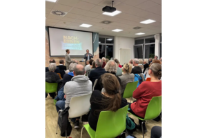 Zwei Männer sprechen vor Publikum in einem hellen Raum mit Projektor und Leinwand